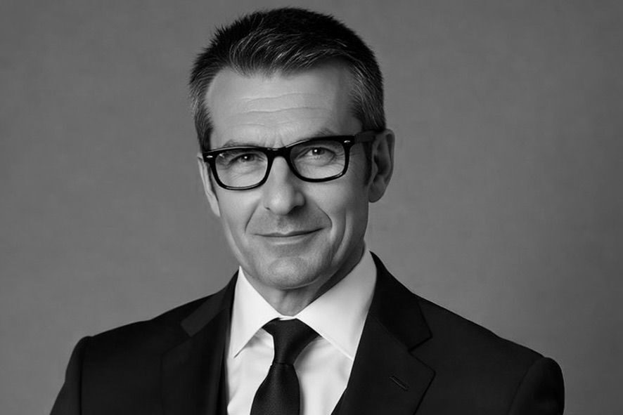 Portrait of a middle-aged man in a black suit, white shirt, black tie, and glasses looking at the camera.