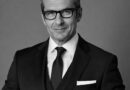 Foto Jose - eVTOL Insights Professional portrait of a middle-aged man in a black suit and tie, wearing glasses against a neutral background.