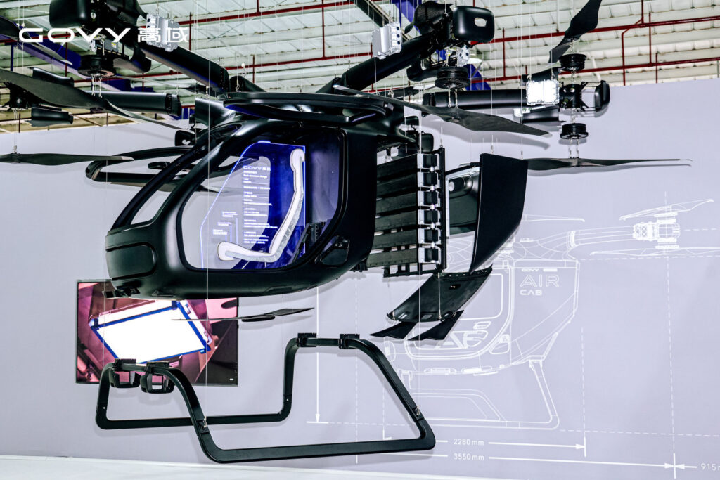 Suspended black helicopter cockpit exhibit with rotor blades overhead and blue-lit cockpit interior in a showroom.