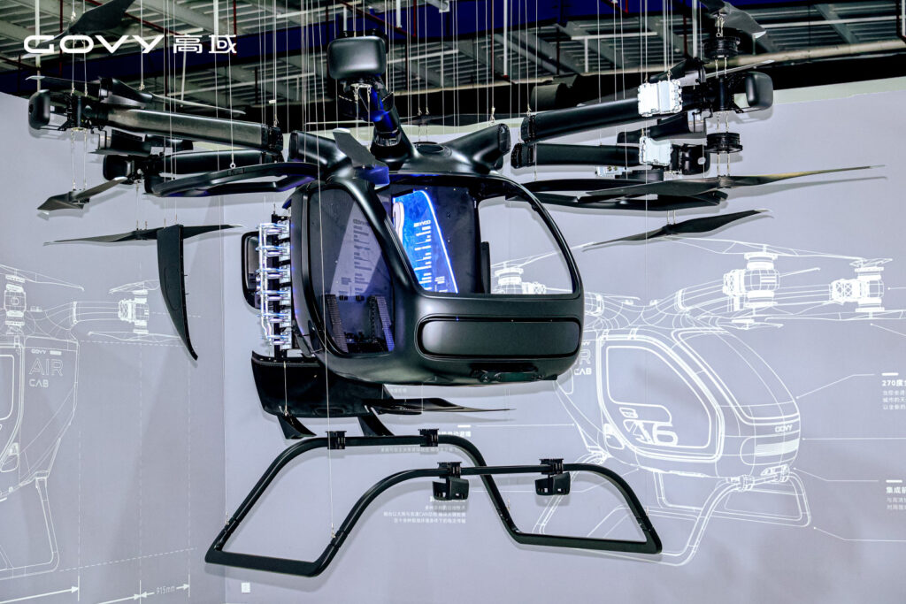 Futuristic helicopter cockpit prototype suspended from the ceiling with exposed rotor arms and a blue-lit cockpit window.