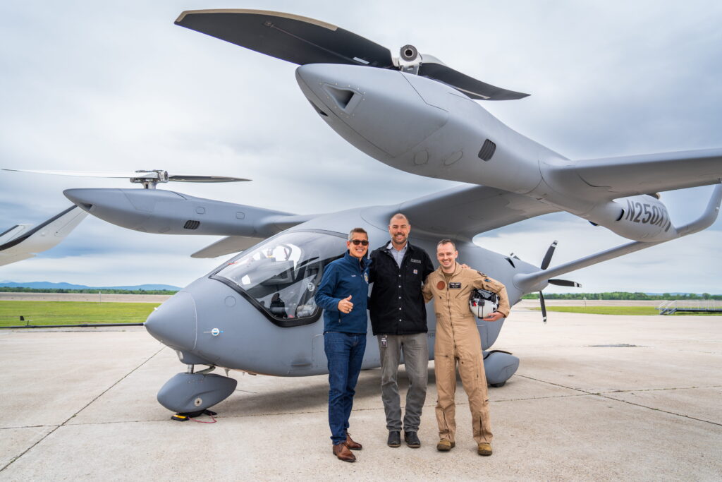 Three people in blue and tan uniforms pose in front of a gray, fan-powered aircraft on a tarmac.