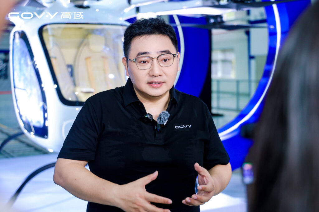 Man with glasses in a black polo explains something, headset mic clipped on, with a white-and-blue helicopter exhibit in the background.