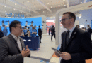 Two businessmen talk at a tech expo with a Joby drone booth and large blue display in the background.