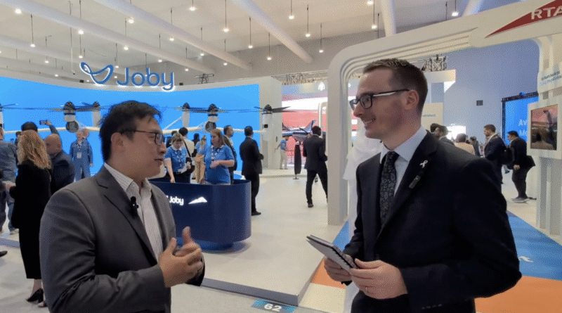 Two businessmen talk at a tech expo with a Joby drone booth and large blue display in the background.