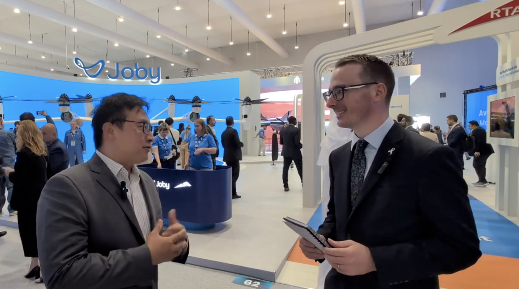 Two businessmen talk at a tech expo with a Joby drone booth and large blue display in the background.