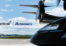 Close-up of a helicopter nose with rotor hub above and the Vertical Astronics logo on the fuselage against a blue sky.