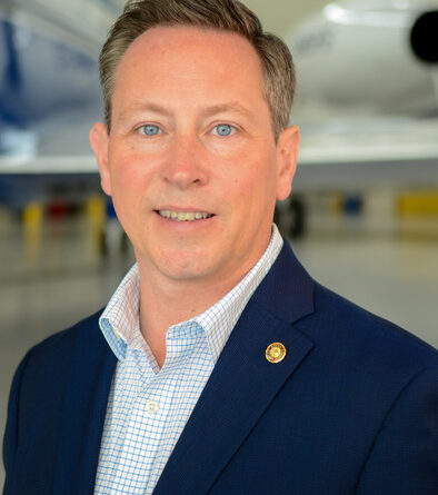 Portrait of a man in a navy blazer and checkered shirt, with a jet in a hangar behind him.