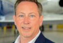 Portrait of a man in a navy blazer and checkered shirt, with a jet in a hangar behind him.