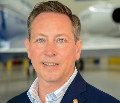 Portrait of a man in a navy blazer and checkered shirt, with a jet in a hangar behind him.