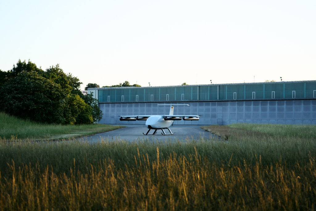 Victor U250 1 - eVTOL Insights Small white drone aircraft with rotor blades parked on a runway in front of a blue hangar, with tall grass in the foreground.