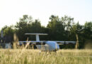 Small white glider aircraft parked on a grassy field with trees in the background and a clear sky above.
