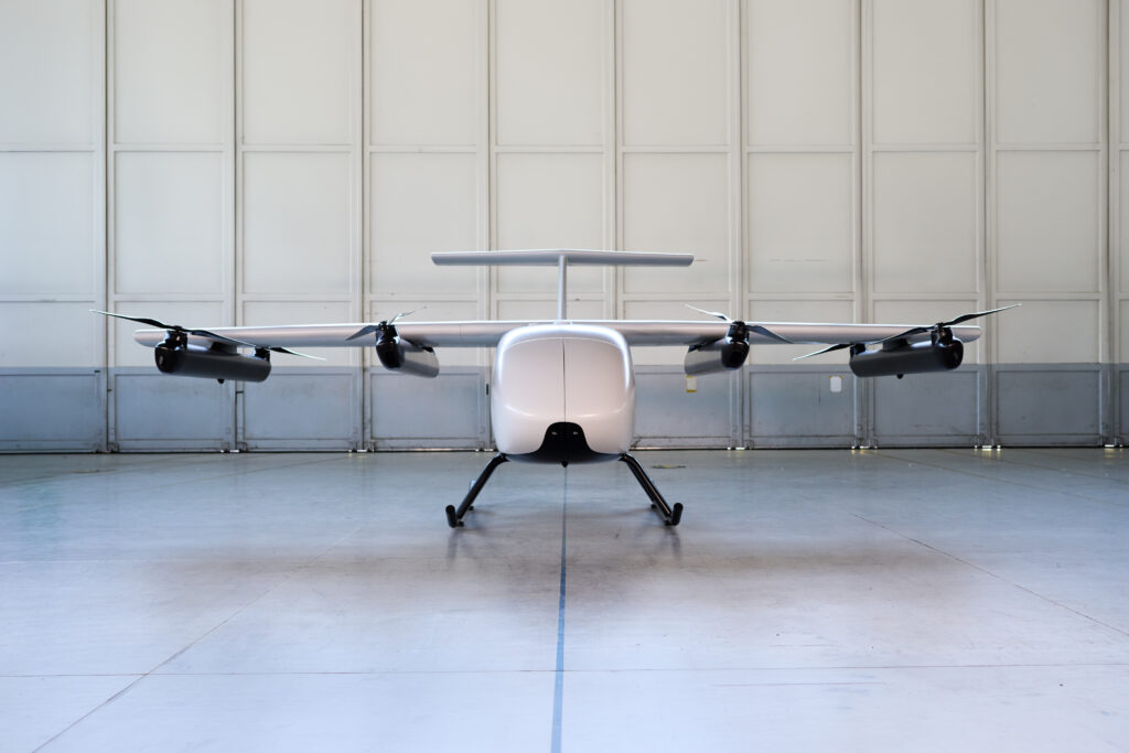 Victor U250 5 - eVTOL Insights Front view of a small white twin-engine airplane parked inside a hangar with tall white doors behind it.