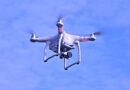 White quadcopter drone with red accents hovering in a clear blue sky, camera mounted underneath