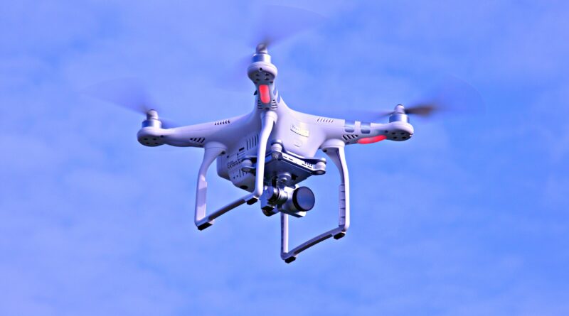 White quadcopter drone with red accents hovering in a clear blue sky, camera mounted underneath