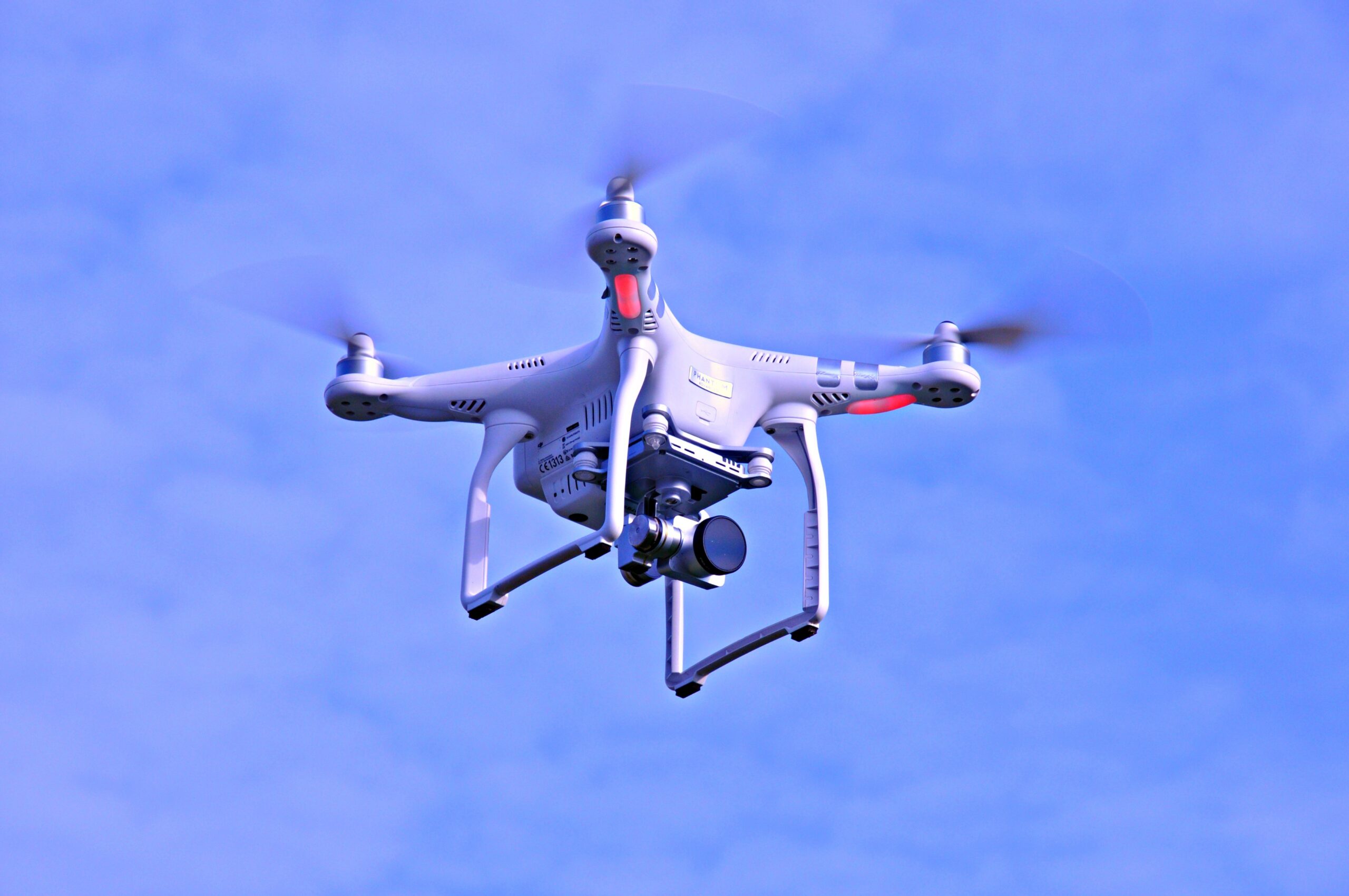 White quadcopter drone with red accents hovering in a clear blue sky, camera mounted underneath