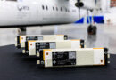 Row of white and orange battery modules on a dark floor with a blurred airplane in the background, suggesting an aircraft hangar setting.
