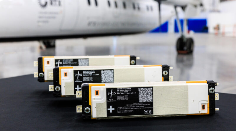 unnamed 51 - eVTOL Insights Row of white and orange battery modules on a dark floor with a blurred airplane in the background, suggesting an aircraft hangar setting.
