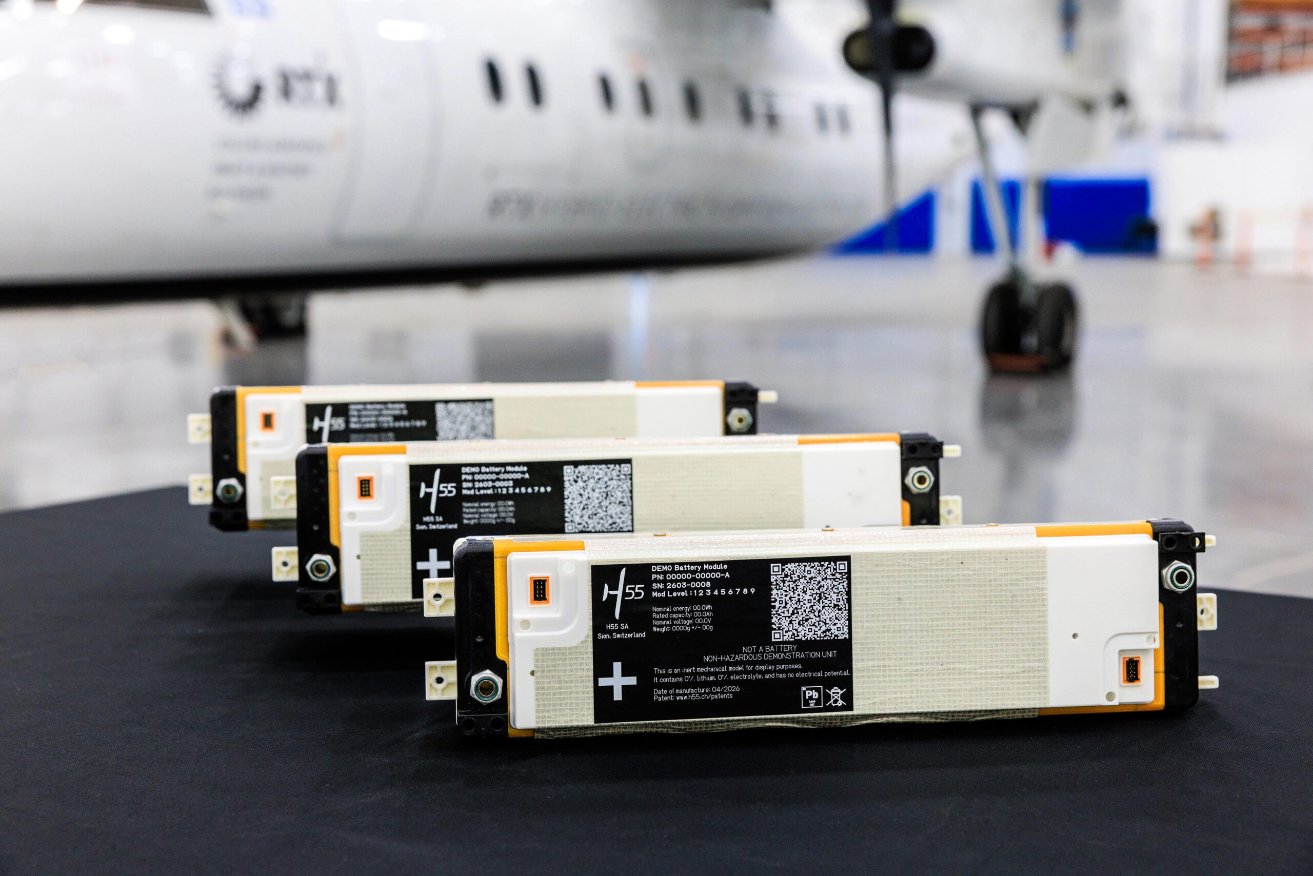 Row of white and orange battery modules on a dark floor with a blurred airplane in the background, suggesting an aircraft hangar setting.