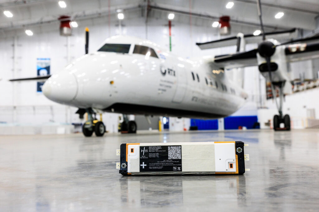 Close-up of a large battery module on the hangar floor with a white airliner parked in the background.