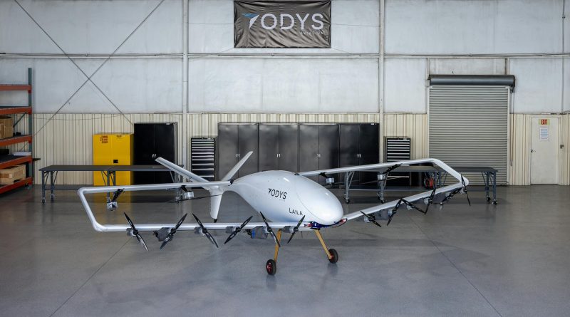 White multi-rotor UAV on a stand in a hangar, ODYS Aviation branding on the fuselage and a banner overhead.