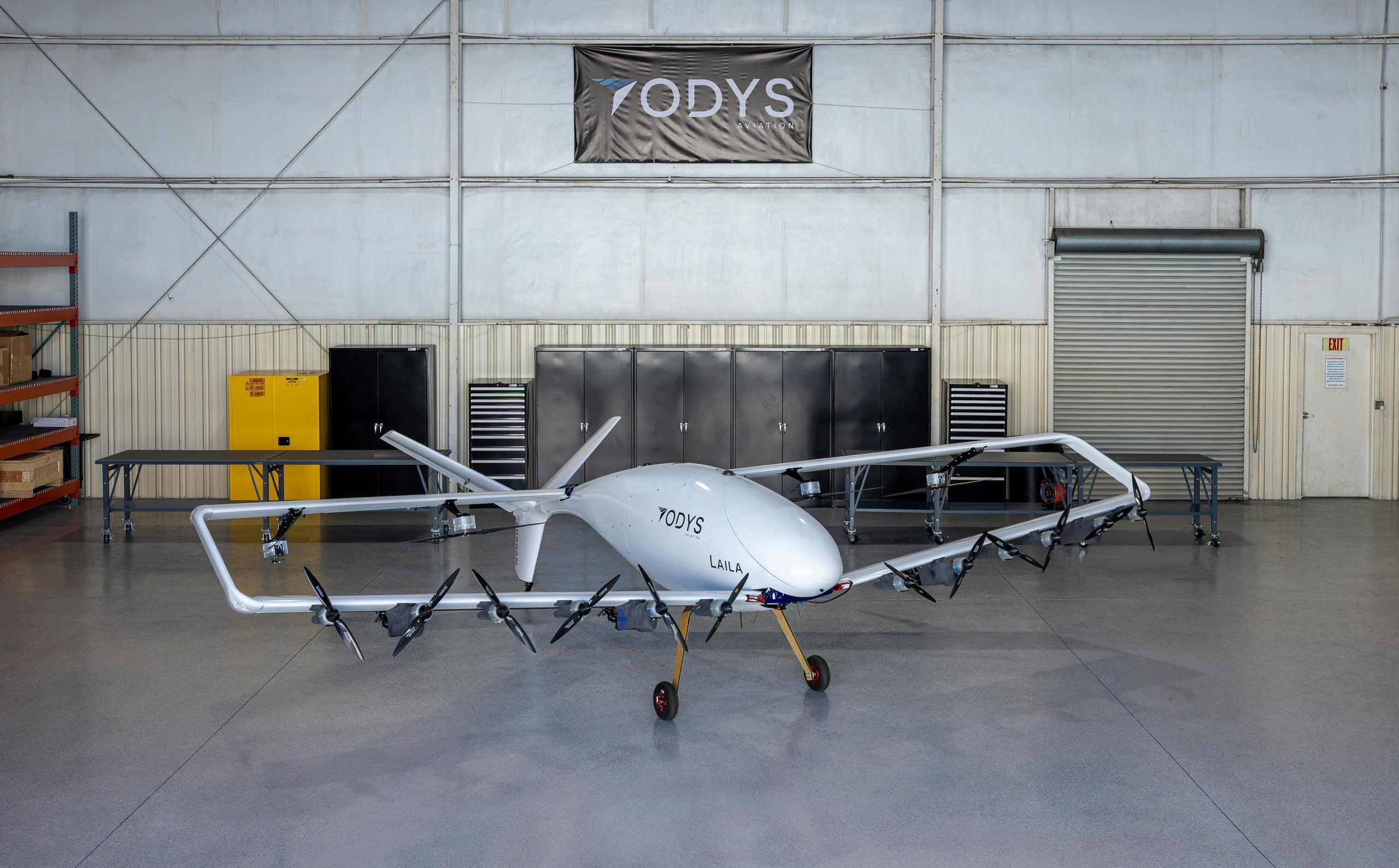 White multi-rotor UAV on a stand in a hangar, ODYS Aviation branding on the fuselage and a banner overhead.