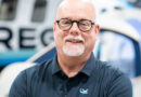 Smiling bald man with round glasses and white goatee, arms crossed in a navy polo in a workshop.