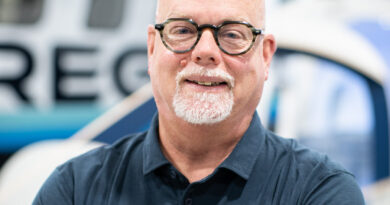 Smiling bald man with round glasses and white goatee, arms crossed in a navy polo in a workshop.