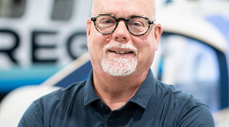 Smiling bald man with round glasses and white goatee, arms crossed in a navy polo in a workshop.