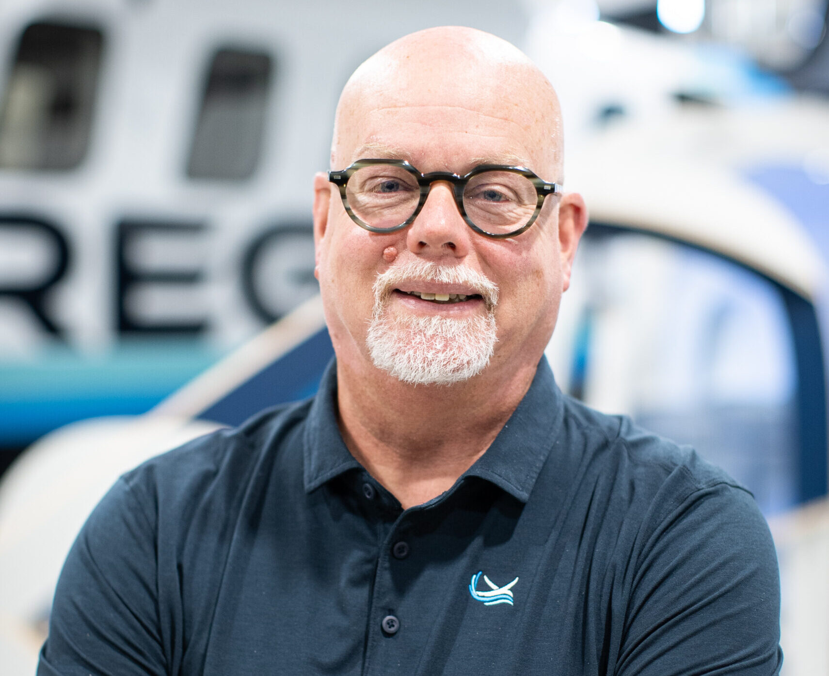Smiling bald man with round glasses and white goatee, arms crossed in a navy polo in a workshop.