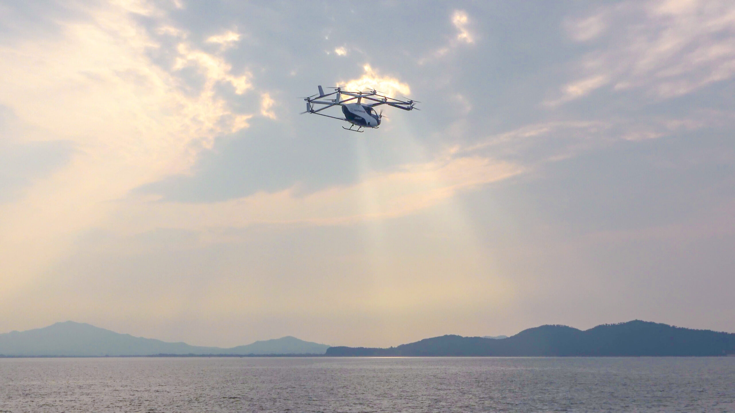 Light helicopter flying above a calm body of water with low hills on the horizon at sunset