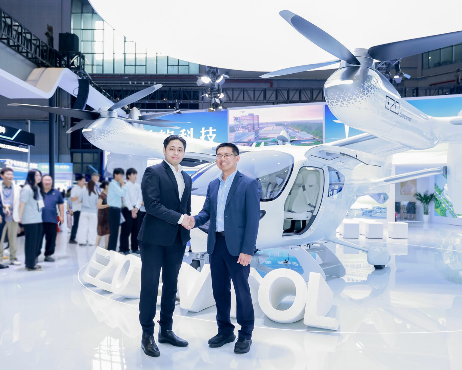 Two businessmen in suits shake hands in front of a white helicopter at an indoor expo, with attendees in the background.