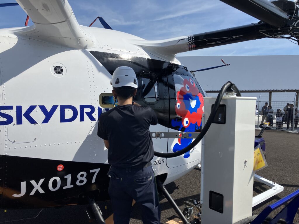 Person in a white helmet standing beside a white helicopter with blue lettering and a red-blue cartoon octopus decal by the cockpit.