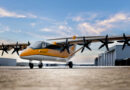 Orange and white multi-propeller aircraft parked on a tarmac beside hangars, under a blue sky.