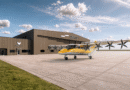 Yellow twin‑engine Electra aircraft parked on a concrete apron beside a brown Signature Aviation hangar under a blue sky.