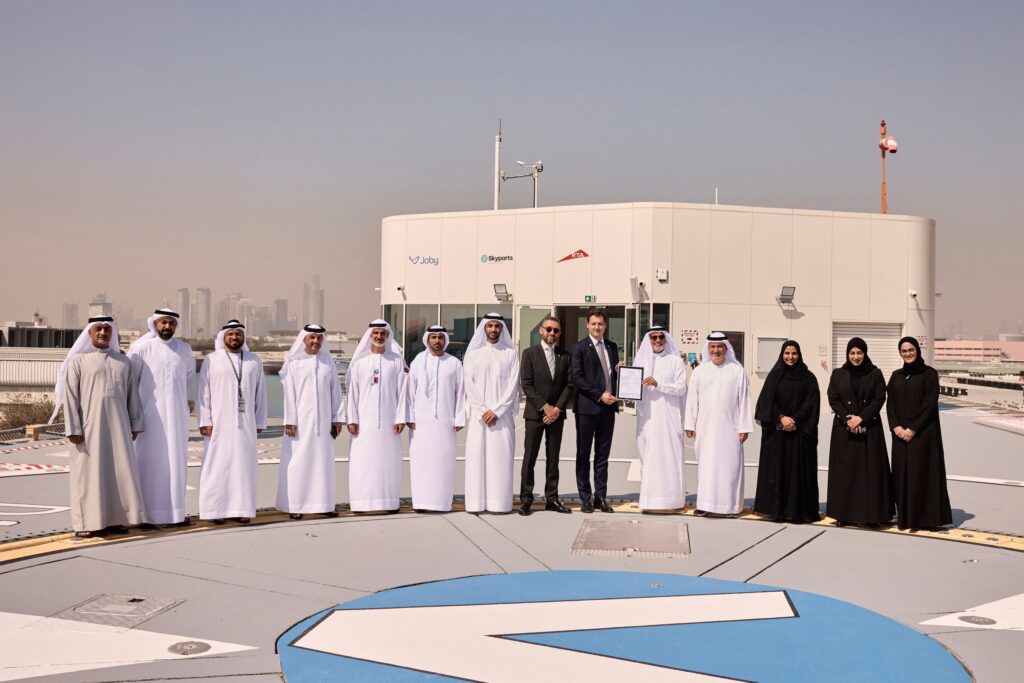 Group of men in white kanduras and women in black abayas, plus a few in suits, posing on a rooftop with a city skyline behind them.