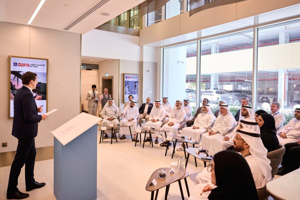 Man in a suit speaks at a lectern to a seated audience in a bright conference room, attendees in white traditional dress nearby