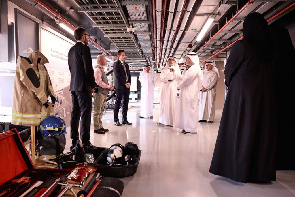 Group of men in traditional white robes and business attire standing in a tech facility, discussing amidst exposed pipes and equipment, with safety gear and helmets laid out in the foreground.