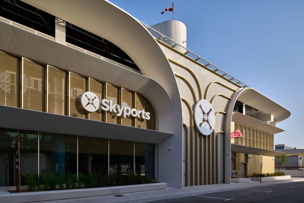 Modern airport-style building facade with curved white arches, gold fins, and the Skyports sign against a clear blue sky.