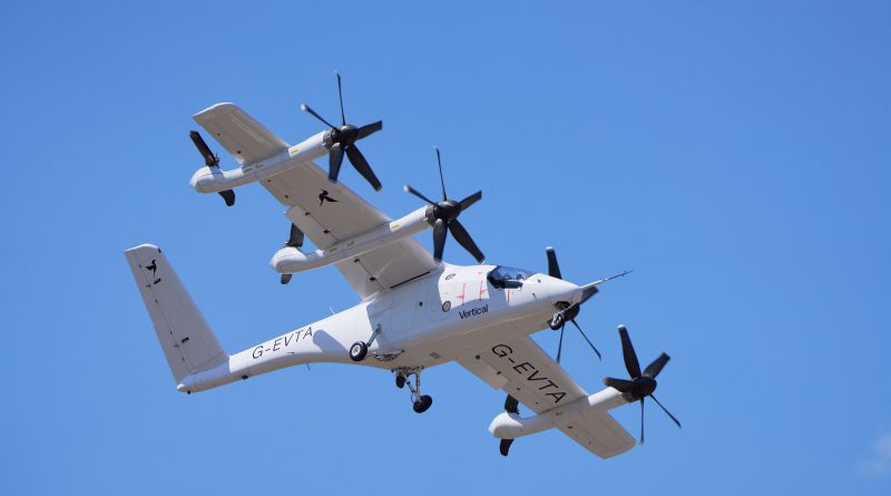 White twin-rotor aircraft in midflight against a clear blue sky, with 'Vertical' branding visible on the fuselage and registration G-EVTA.