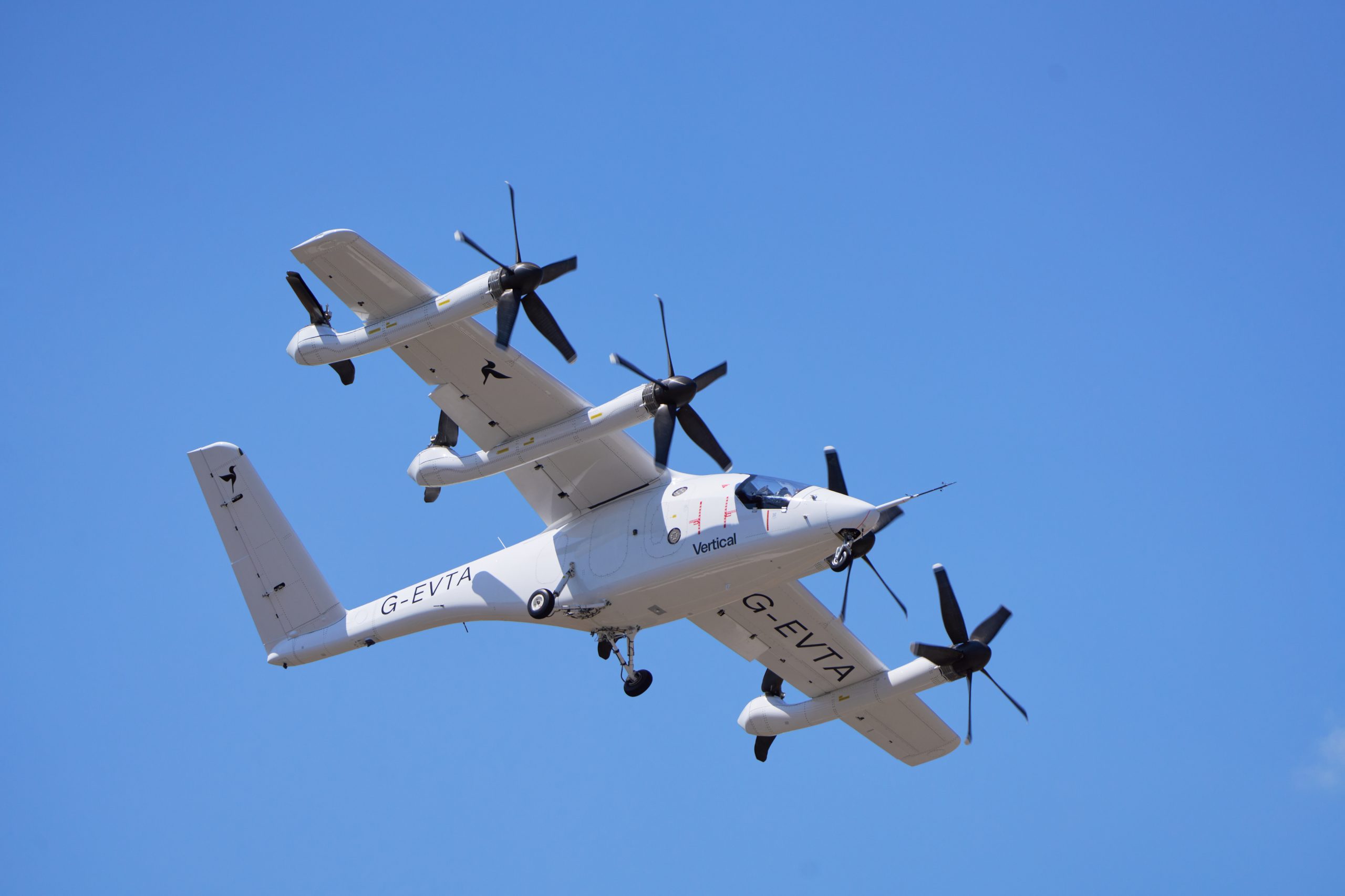 White twin-rotor aircraft in midflight against a clear blue sky, with 'Vertical' branding visible on the fuselage and registration G-EVTA.