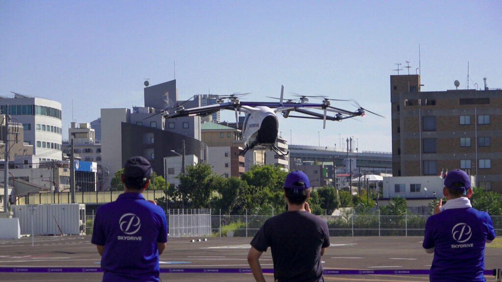 Large multi-rotor drone hovering above a fenced urban area as three observers in purple SKYDIVE shirts watch.