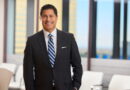 Smiling businessman in a dark suit and striped tie standing in a bright office lobby with glass walls behind him.