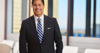 Smiling businessman in a dark suit and striped tie standing in a bright office lobby with glass walls behind him.