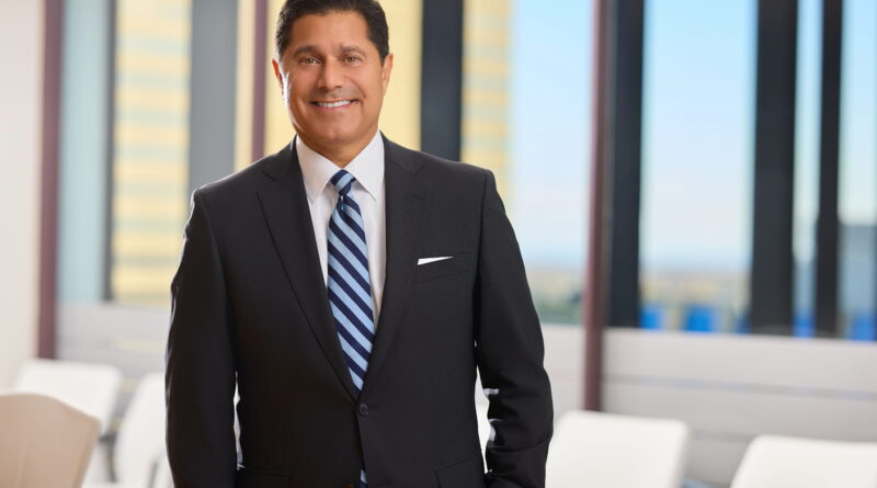 Smiling businessman in a dark suit and striped tie standing in a bright office lobby with glass walls behind him.