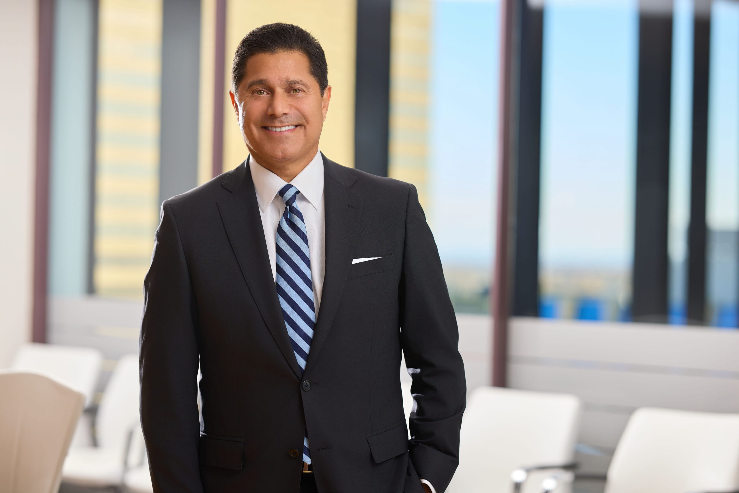 Smiling businessman in a dark suit and striped tie standing in a bright office lobby with glass walls behind him.
