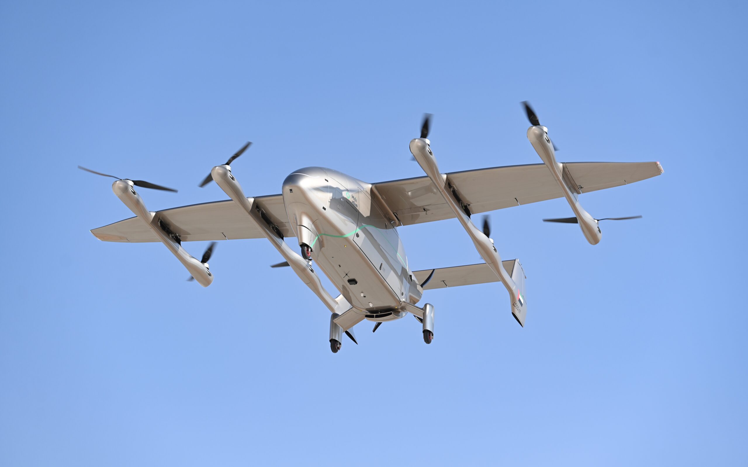 Tilt-rotor aircraft in flight viewed from below, four propellers spinning and landing gear visible against a clear blue sky
