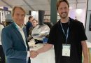 Two men shake hands at a trade show, smiling, with badges and a large machine in the background.