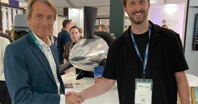 Ludens-Dovetail 21-07-2026 - eVTOL Insights Two men shake hands at a trade show, smiling, with badges and a large machine in the background.