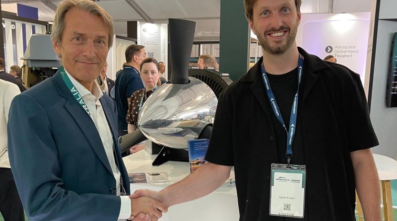Ludens-Dovetail 21-07-2026 - eVTOL Insights Two men shake hands at a trade show, smiling, with badges and a large machine in the background.
