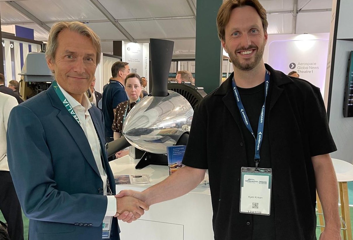 Two men shake hands at a trade show, smiling, with badges and a large machine in the background.
